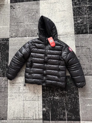 Svart dunjacka från Canada Goose - Säljer en svart dunjacka från Canada Goose med huva och dragkedja. Jackan har två sidofickor med dragkedja och klassisk logotyp på vänster ärm. Perfekt för kalla vinterdagar och riktigt snygg streetstil.