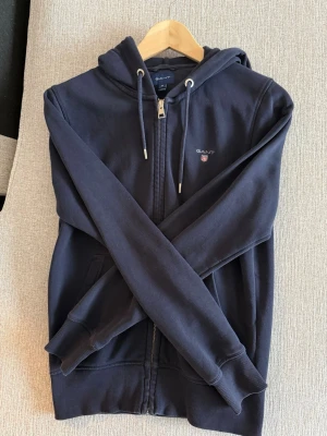 Mörkblå zip hoodie från GANT - Snygg mörkblå hoodie från GANT med dragkedja framtill, huva med dragsko och fickor på sidorna. Klassisk broderad GANT-logga på bröstet. Tillverkad i mjuk bomullsblandning som känns skön mot huden. Perfekt för en chill och avslappnad stil.