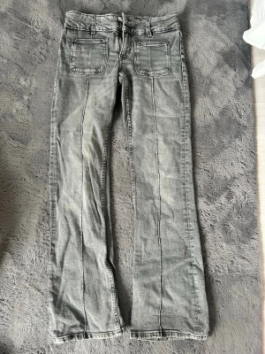 Grå bootcut jeans med framfickor - Säljer ett par grå bootcut jeans och markerade sömmar framtill. Jeansen har två stora framfickor och två bakfickor. Klassisk femficksmodell med knapp och dragkedja. Materialet är denim i bomull. Perfekta för en avslappnad och trendig look.