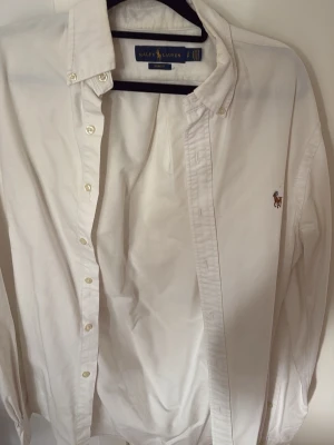 Vit skjorta från Ralph Lauren - Klassisk vit skjorta från Ralph Lauren med broderad logga på bröstet. Skjortan har knappar framtill, krage och långa ärmar. Tillverkad i bomull och passar perfekt till en stilren look.