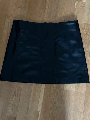 Svart kort kjol i skinnimitation - Snygg svart minikjol i skinnimitation med enkel design och diskret dragkedja bak. Kjolen har en rak passform och är perfekt för att skapa en edgy look. Passar till både t-shirt och stickad tröja för en trendig outfit.