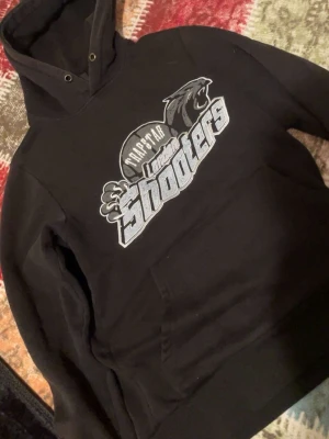 Svart Trapstar hoodie med tryck - Säljer en svart hoodie från Trapstar med stort vitt och grått tryck på bröstet där det står 'London Shooters' och en pantergrafik. Hoodien har huva med dragsko och en klassisk känguruficka framtill. Perfekt för dig som gillar streetwear och vill sticka ut.