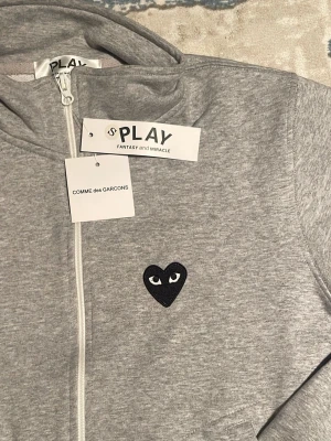 Grå hoodie från Comme des Garçons Play - Säljer en grå hoodie från Comme des Garçons Play med dragkedja framtill och det ikoniska svarta hjärtat med ögon broderat på bröstet. Tillverkad i mjukt bomullsmaterial och har ribbade muddar vid ärmslut och nederkant. Perfekt för en chill och stilren look.