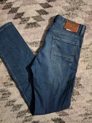 Blå jeans från Tommy Hilfiger - Säljer ett par klassiska blå jeans från Tommy Hilfiger med rak passform och snygga detaljer som läderpatch bak och den ikoniska loggan på bakfickan. Jeansen är i denim och har kontrastsömmar samt en liten tygdetalj i grönt, vitt och rött.