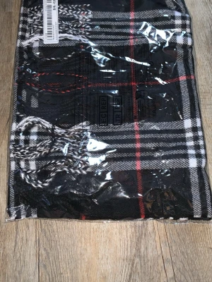 Svart-rutig halsduk med fransar - Snygg halsduk i svart med vitt och rött rutmönster. Klassisk design med fransar i ändarna, perfekt för att lyfta din outfit. Tillverkad i mjukt tyg som känns skönt mot huden. Passar dig som gillar stilrena accessoarer med en twist.
