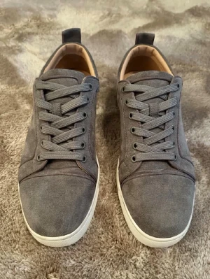 Gråa Louboutin sneakers i mocka - Snygga mock distincta sneakers från Christian Louboutin i grått med klassisk röd sula och vita kanter. Skorna har rund tå, gråa skosnören och diskret branding på insidan. Perfekta för dig som vill ha en clean och exklusiv look.