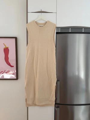 Beige stickad ärmlös klänning - Beige stickad klänning i mjukt material med ribbade kanter och slits på sidan. Ärmlös modell med rund halsringning, perfekt för lager på lager eller sommarlooken. Enkel och stilren design som passar till många olika stilar.