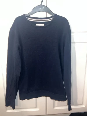 Mörkblå stickad tröja - En enkel och stilren mörkblå stickad tröja. Tröjan har rund halsringning, långa ärmar och ribbade muddar vid ärmslut och nederkant. Perfekt att matcha med jeans eller kjol för en clean look. Passar bra över en skjorta.