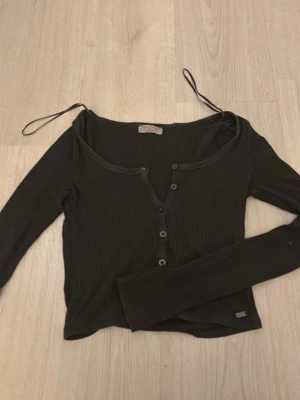 Svart ribbad topp med knappar - Trendig svart ribbad topp med lång ärm och knappar framtill. Toppen har en croppad passform och tunna axelband som ger en unik look. 