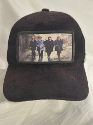 Svart truckerkeps Maggiore med print - Svart truckerkeps från Maggiore med mesh på sidorna och bak, samt ett stort tygmärke framtill med motiv av fyra personer i kostym. Kepsen har justerbar snapback och en liten metallogo på sidan. Perfekt accessoar för en cool streetstil.