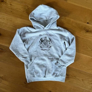 Grå hoodie från BVALDI  - Snygg grå hoodie från BVALDI med stort svart tryck på bröstet som visar ett vapensköldmotiv och texten 'Côte d’Azur Club'. Hoodien har huva och magficka, samt ribbadeMuddar vid ärmslut och nederkant. Perfekt för en chill och stilren look. Nypris är 999kr och den är som ny 