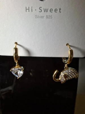 Guldörhängen med hjärta och elefant - Unika örhängen i guldton från Hi Sweet, tillverkade i silver 925. Ena örhänget har ett gnistrande hjärta i klar sten och det andra ett elefantformat hänge dekorerat med små stenar. Perfekt för dig som gillar lekfulla och glittriga accessoarer.