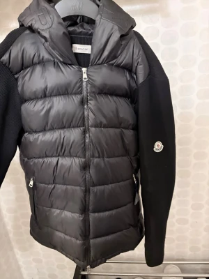 Moncler jacka - Snygg svart pufferjacka från Moncler med quiltad front och rygg i glansigt material, stickade ärmar och huva. Jackan har dragkedja framtill, två fickor med dragkedja och Moncler-logga på ärmen. Perfekt för kalla vinterdagar.