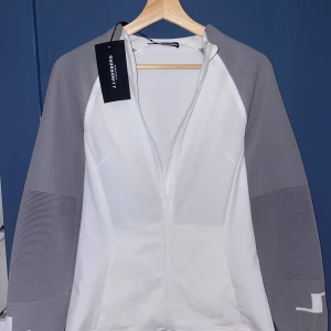 Vit och grå vindjacka J.Lindeberg - Snygg vit och grå dam vindjacka från J.Lindeberg med hel dragkedja framtill. Jackan har ribbade grå ärmar och vita detaljer, samt diskret logga på ärmen. 