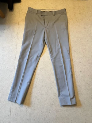 Kostymbyxa-dressmann - Snygga beige kostymbyxor med klassiskt pressveck och bälteshällor. Tillverkade i en mix av polyester, bomull och elastan för skön passform och stilren look. Perfekta till skjorta eller kavaj för en clean outfit.