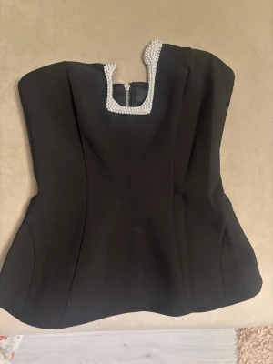 Svart korsettopp med pärldetaljer Zara - Stilren svart korsettopp från Zara med fyrkantig urringning dekorerad med pärlor. Toppen är axelbandslös och har figurnära passform med dragkedja bak. Perfekt för dig som vill ha en elegant och trendig look.