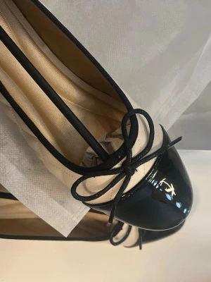 Beige och svarta ballerinaskor - Stilrena ballerinaskor i beige quiltat material med svart lackad tå och svart kantband. Dekorativ rosett framtill ger en klassisk look. Perfekta för dig som gillar en chic och tidlös stil.