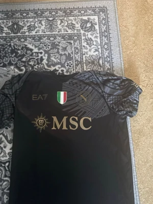 Svart Napoli matchtröja EA7 - Snygg svart matchtröja från Napoli med guldfärgade detaljer och loggor. Tröjan har geometriskt mönstrade ärmar, klubbmärke och sponsortryck på bröstet samt tryck på ryggen. Tillverkad i ett lätt och funktionellt material som passar perfekt till träning eller match.