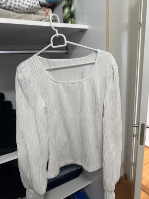 Vit långärmad blus med struktur - Säljer en vit blus med lång ärm och snygg struktur i tyget. Blusen har en bred, rundad urringning och är tillverkad i ett lätt, vågigt material som ger en trendig look. Perfekt att styla med jeans eller kjol för en fräsch outfit.