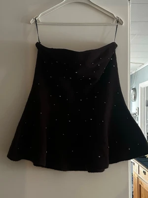 Svart axelbandslös tubtopp med pärlor - Snygg mörkbrun axelbandslös tubtopp med pärldetaljer. Toppen har en lätt utsvängd form och tunna axelband som kan tas bort för en axelbandslös look. Perfekt för dig som vill ha en stilren men ändå unik topp. ( obs ser mörkare ut på bild än vad den är)