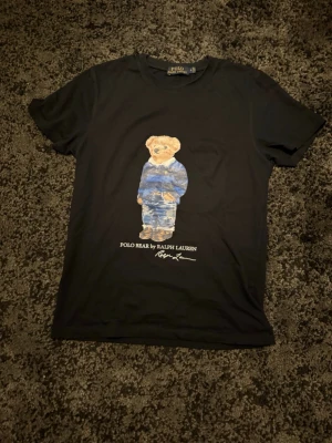 Svart Polo Bear t-shirt från Ralph Lauren - Snygg svart t-shirt från Ralph Lauren med ikoniskt Polo Bear-tryck på bröstet. T-shirten har rund hals och korta ärmar. Tillverkad i mjuk bomull som känns skön mot huden. 
