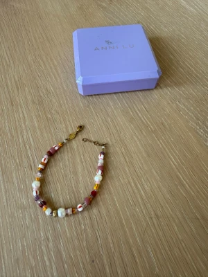 Färgglatt pärlarmband från Anni Lu - Säljer ett färgglatt armband från Anni Lu med mixade pärlor i vitt, rosa, orange och guldtoner. Armbandet har oregelbundna former på pärlorna och guldfärgade metalldetaljer. Perfekt accessoar för att addera färg till din stil!