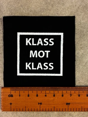 Svart tygmärke KLASS MOT KLASS - Säljer ett svart tygmärke med vit screen print och texten 'KLASS MOT KLASS' i versaler, inramad av en vit fyrkant. Märket är fyrkantigt och passar perfekt att sy eller stryka på jackor, väskor eller kepsar för en personlig touch.