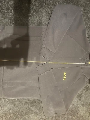 Svart zip-hoodie från BOSS - Svart hoodie med dragkedja från BOSS. Gult BOSS-tryck på bröstet, gröna detaljer på insidan av huvan och två fickor framtill. Klassisk passform med ribbade muddar och huva. Perfekt för en stilren och avslappnad look.