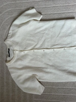 Vit kortärmad kofta från 157 - Stilren vit kofta från 157 med korta ärmar och rund hals. Koftan har ribbade kanter och knäpps med små pärlknappar framtill. Perfekt att matcha med jeans eller kjol för en clean look.