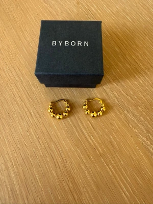 Guldiga örhängen från Byborn - Snygga örhängen i guldfärg från Byborn. De är runda hoops dekorerade med små pärlliknande detaljer som ger en trendig och elegant look. Perfekta för att lyfta din stil med en touch av guld.