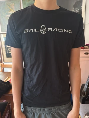 Svart Sail Racing t-shirt - Snygg svart t-shirt från Sail Racing med vit logga tryckt över bröstet. Klassisk rund halsringning och korta ärmar. Perfekt till jeans eller shorts för en clean och sportig look. Tillverkad i mjuk bomull för skön känsla hela dagen.