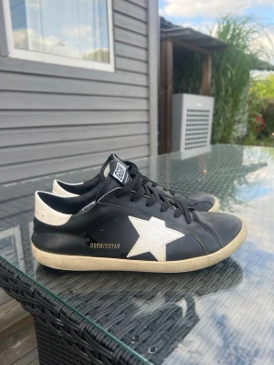 Svarta GGDB sneakers med vit stjärna - Snygga svarta sneakers från Golden Goose med vit stjärna på sidan och vit häl. Skorna har svarta skosnören, GGDB-logga på plösen och en klassisk låg siluett. Ovandelen är i läder och sulan är beige för en cool kontrast.