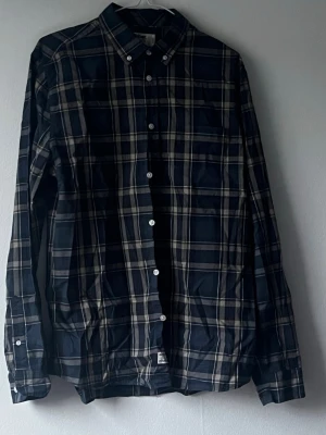 Mörkblå rutig skjorta med knappar - Säljer en mörkblå skjorta med rutigt mönster i beige och vitt. Skjortan har långa ärmar, klassisk krage och knäppning framtill. Perfekt för dig som gillar en stilren och tidlös look.