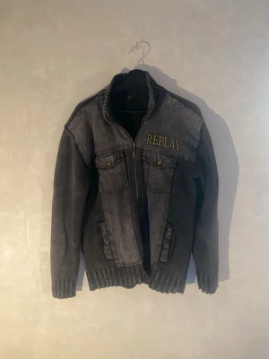 Mörkgrå jeansjacka från Replay - Snygg mörkgrå jeansjacka från Replay med dragkedja, ribbade muddar och hög krage. Jackan har två bröstfickor med knappar och två sidofickor. Stora Replay-loggan broderad på bröstet ger en cool touch. Perfekt för dig som gillar streetwear.