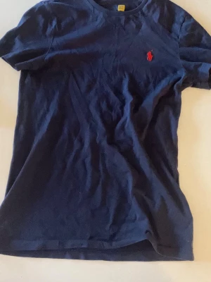 RALPH LAUREN T-SHIRT - Snygg mörkblå Ralph Lauren t-shirt med klassisk passform och en liten röd logga broderad på bröstet. Tillverkad i mjuk bomull och har korta ärmar. Perfekt till jeans eller shorts för en enkel och stilren look.
