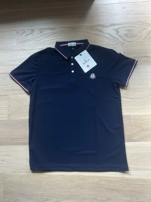 Mörkblå moncler pikétröja - Snygg mörkblå pikétröja med vita knappar och tunna röda och vita ränder på krage och ärmslut. Tröjan har en klassisk passform och ett broderat märke på bröstet. Perfekt till jeans eller shorts för en clean look.