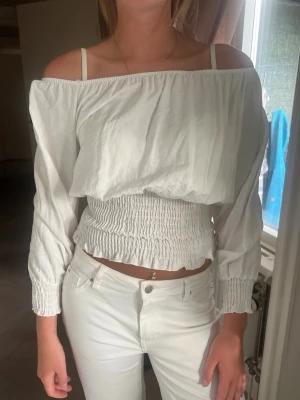 Vit offshoulder blus med smock - Trendig vit offshoulder blus med långa ärmar och smockad midja. Toppen har en luftig passform upptill och sitter tightare över magen. Perfekt att styla med höga jeans eller kjol för en fräsch look.