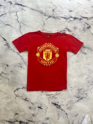 Manchester United T-shirt  - En mycket fin Manchester United T-shirt i barn storlek. Plagget har inte använts utan bara legat i källaren. Det finns små hål ( kolla de sista bilderna, de är två bilder p en defekt ) men de syns absolut inte både för de är så små men främst för att de är på sidan.                      Priset kan diskuteras!
