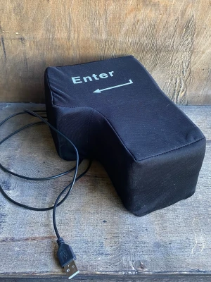 Stor USB Enter-knapp kudde - En stor svart Enter-knapp i tygmaterial som ansluts via USB. Perfekt som stresskudde eller rolig datorpryl på skrivbordet. Formad som en klassisk Enter-tangent med vit text och pil. Passar till datorer för extra knapptryck.