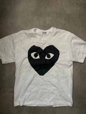 Vit Comme des Garçons Play t-shirt - Vit t-shirt från Comme des Garçons Play med ett stort svart hjärta med ögon tryckt på bröstet. Klassisk rund hals och korta ärmar. Tillverkad i mjuk bomull för en skön och avslappnad stil.