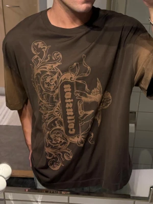 Brun t-shirt med tryck från Collusion - Säljer en brun t-shirt från Collusion med stort grafiskt tryck i beige på framsidan. T-shirten har korta ärmar och en relaxed passform. Materialet är mjuk bomull och trycket har en vintage vibe med ängel och ornament.