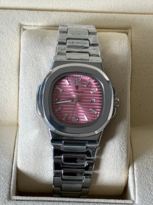 Silverfärgad klocka med rosa urtavla - Stilren klocka från Peodagar med silverfärgad länkarmband i metall och en rosa urtavla med randigt mönster. Urtavlan har datumvisning och markerade index med stenar vid vissa timmar. Perfekt accessoar för dig som vill sticka ut.