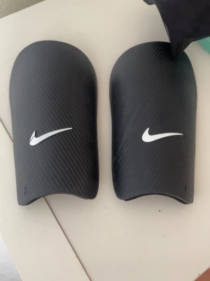 Nike benskydd - Ett par Nike benskydd i svart färg. Benskydden kommer med en förvaringspåse. Klippte av kardborren så man har dom som slip on. Bra skick annars, ingen spricka. Kan fixa mått om det behövs.