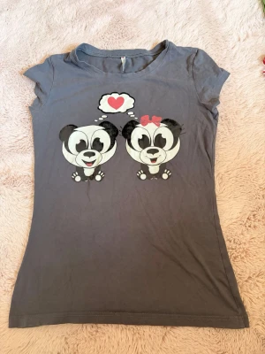 Grå t-shirt med panda-tryck - Söt grå t-shirt med tryck av två pandor på framsidan, där den ena har en röd rosett och ett hjärta i en tankebubbla. T-shirten har rund hals och korta ärmar. Perfekt för dig som gillar gulliga motiv och vill ha en bekväm vardagströja eller pyjamas tröja💗