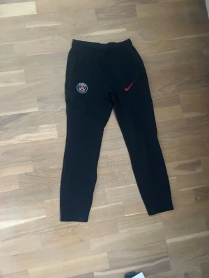 Svarta Nike PSG Dri-FIT träningsbyxor - Säljer ett par svarta träningsbyxor från Nike med PSG-logga och röd Nike Swoosh på vänster ben. Byxorna är i Dri-FIT-material som andas och har resår i midjan samt smal passform. Perfekta för fotbollsträning eller gymmet. Hör av er vid frågor 👍