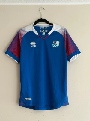 Blå Island fotbollströja Errea VM 2018 - Säljer en blå officiell Island fotbollströja från Errea med KSÍ-logga på bröstet och texten 'FYRIR ISLAND' i nacken. Tröjan har vita och röda detaljer på axlarna och ärmarna samt islandsk flagga på ryggen. Perfekt för match eller träning.