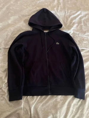 Mörkblå hoodie från Lacoste - Snygg mörkblå hoodie från Lacoste med dragkedja framtill och klassisk krokodil-logga på bröstet. Hoodien har huva med dragsko och två fickor framtill. Perfekt för en avslappnad och stilren look.