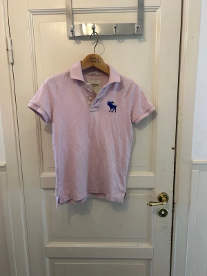 Ljuslila pikétröja från Abercrombie - Snygg ljuslila pikétröja från Abercrombie med klassisk krage, korta ärmar och två knappar framtill. Tröjan har en blå broderad älg på bröstet och är tillverkad i bomull. Perfekt för en avslappnad och stilren look.