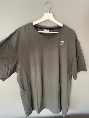 Grå Gymshark t-shirt i bomull XL - Snygg grå t-shirt från Gymshark i storlek XL. Tillverkad i mjuk bomull med klassisk rund hals och korta ärmar. Diskret Gymshark-logga på bröstet. Perfekt för dig som gillar enkel och stilren design.