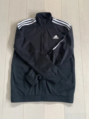 Svart Adidas träningströja - Snygg svart träningströja från Adidas med dragkedja framtill och klassiska vita ränder på axlarna. Tröjan har hög krage, muddar vid ärmslut och nederkant samt Adidas-logga på bröstet. Tillverkad i ett mjukt och stretchigt material som passar perfekt till träning eller chill.